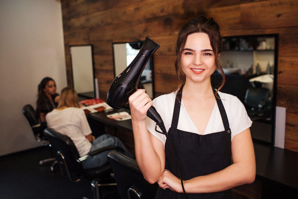 Portret of smiling hairdresser in beauty salon. Beautiful woman in black apron looking at camera and holding professional black hair dryer. Beauty and people concept