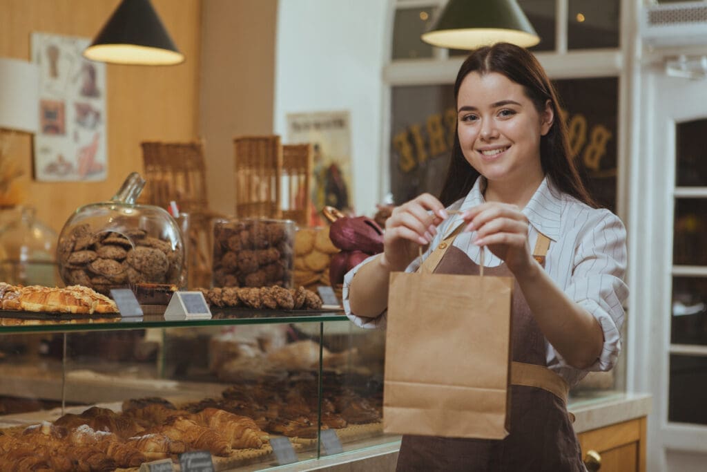 Beautiful cheerful female baker smiling, holding out paper shopping bag to the camera, working at her cafe. Lovely female confectioner selling delicous food at the bakery, copy space
